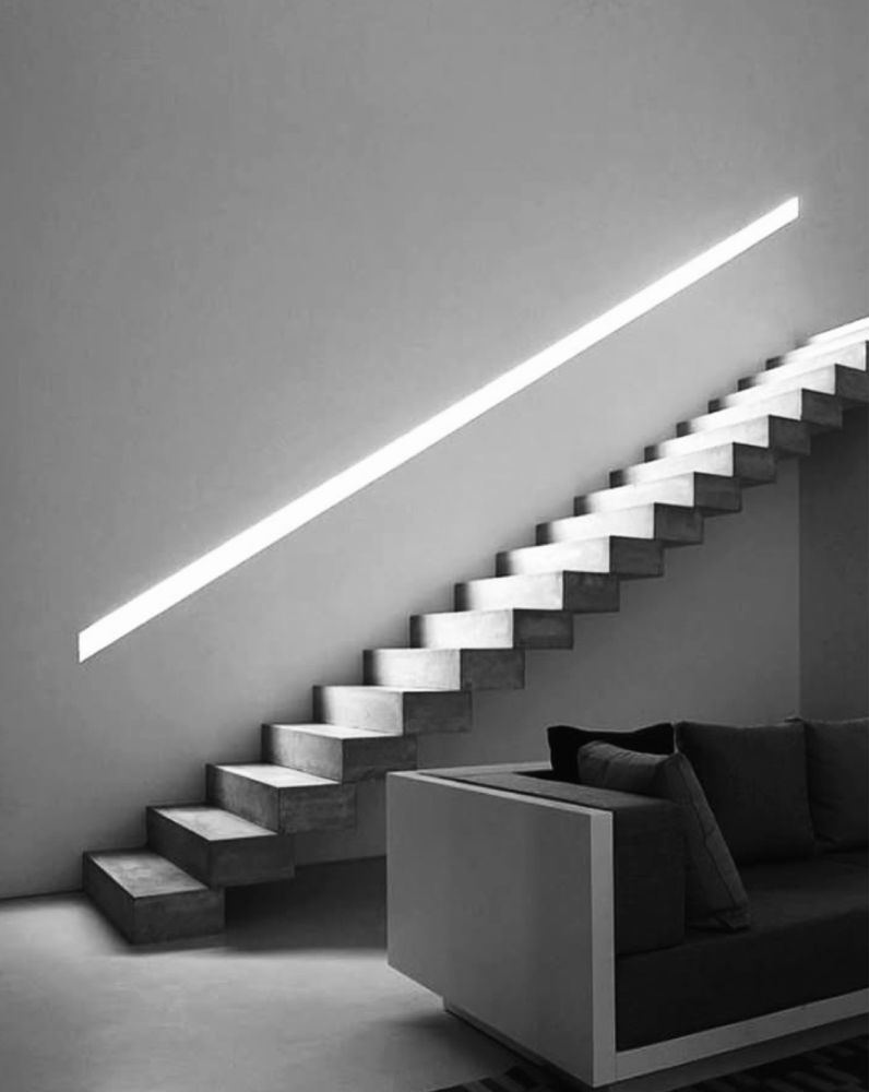 A long straight stairway appears to float off a tall plain wall. Just above it a long angled cutout acts as a recessed handrail and lighting for the stairs using hidden LEDs. 
In the foreground, part of a sofa could indicate the seating area of a contemporary residence.
