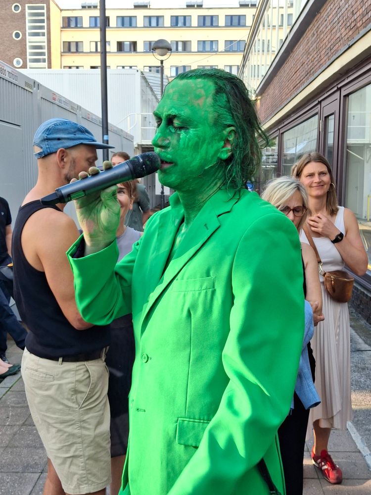 Lars Eidinger im grünen Anzug, grün bemalt und mit Mikrofon in der Hand.