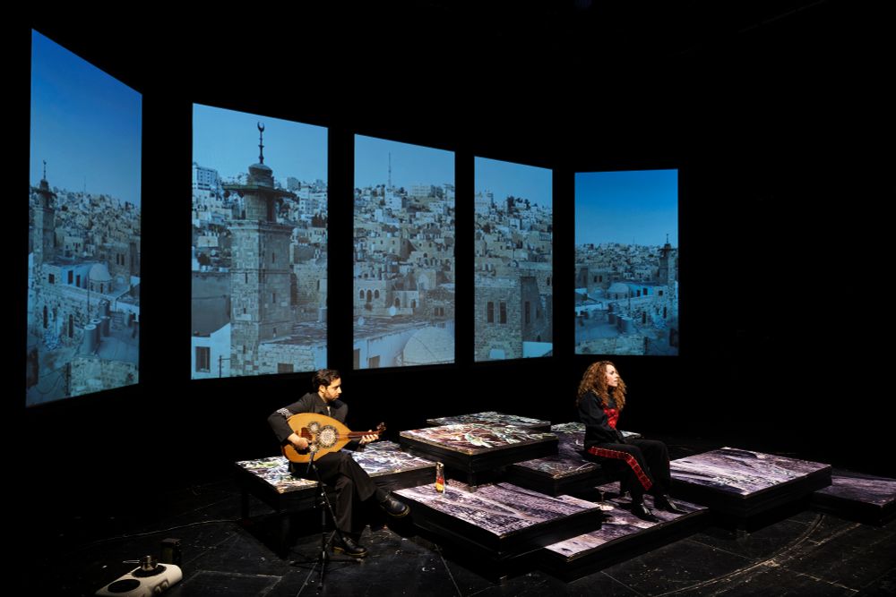 Szenenfoto mit zwei Personen auf einer Bühne aus verschiedenen Podesten, im Hintergrund eine Projektion aus einer Stadtansicht mit Häusern auf fünf Panelen