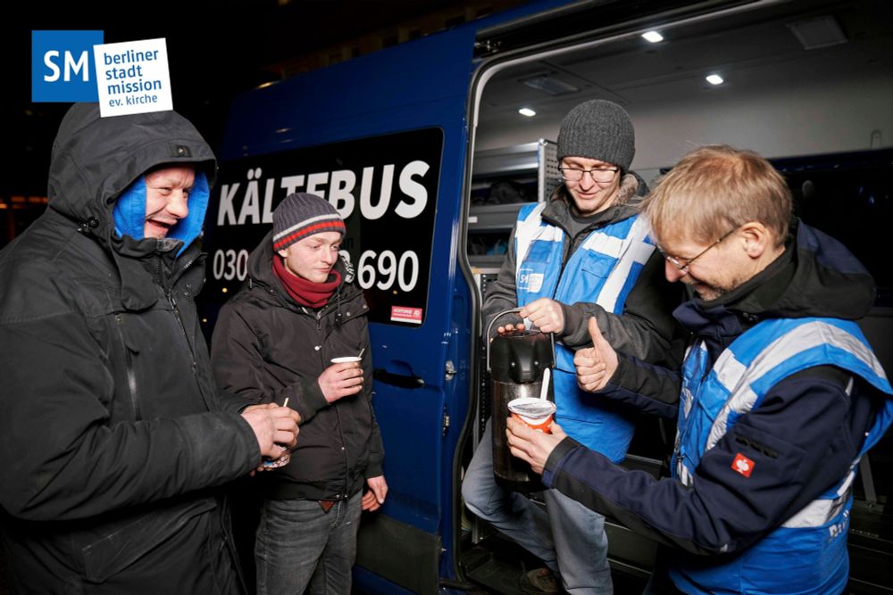 Foto von vier Personen vor dem Kältebus, zwei haben einen Becher mit Heißgetränk, zwei gießen ein Getränk aus einer Thermoskanne in einen Becher