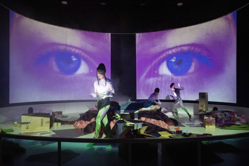 Szenenfoto mit drei Personen in einer runden Arena voller Boxen, im Hintergrund ein großes Augenpaar auf einer Projektion