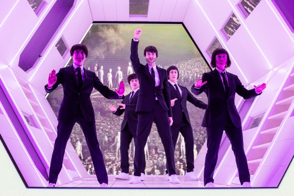 Szenenfoto "RCE" mit fünf Personen in Anzügen und dunklen Perücken in einem tunnelartigen Bühnenbild in violettem Licht