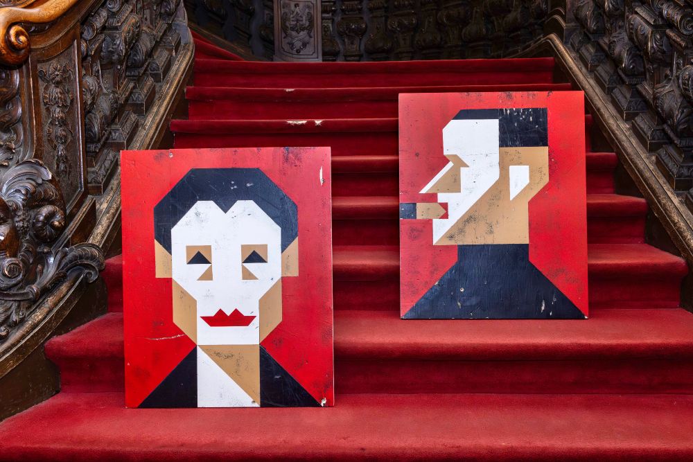 Zwei grafische Porträts von Helene Weigel und Bertolt Brecht in rot, weiß und beige-Farben auf Holzplatten, die auf einer Treppe im BE-Foyer stehen