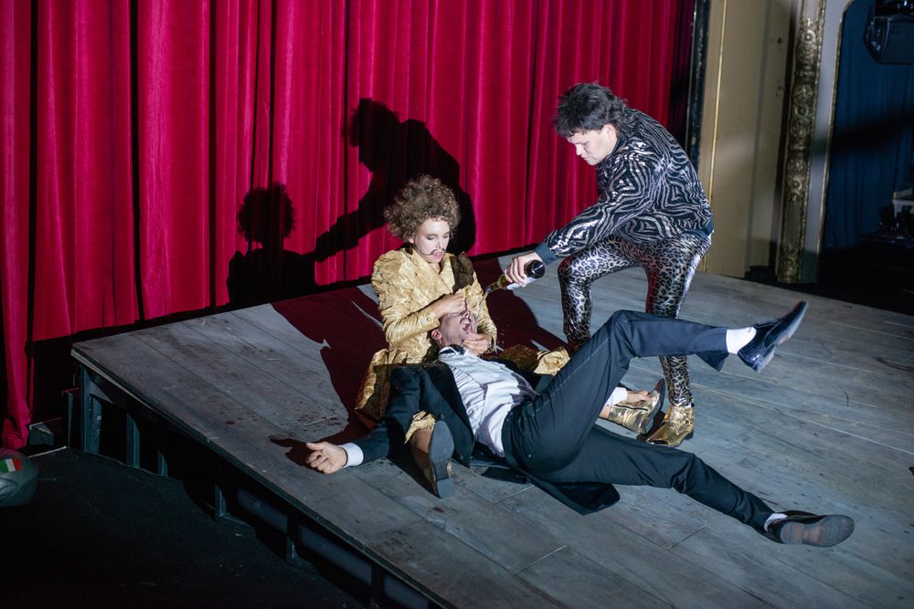 Szenenfoto "Felix Krull", zwei Personen schütten einer weiteren Person auf dem Boden Sekt in den Mund