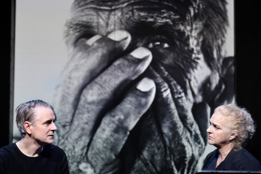 Szenenfoto mit zwei Personen, hinter ihnen eine große Schwarz-Weiß-Projektion eines Fotos einer Person, die beide Hände vors Gesicht hält