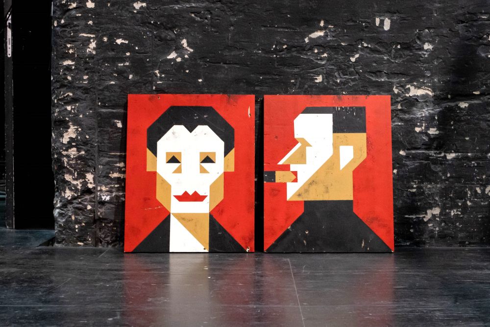 Zwei grafische Porträts von Helene Weigel und Bertolt Brecht in rot, weiß und beige-Farben auf Holzplatten, die an eine schwarze Wand auf der Bühne gelehnt sind