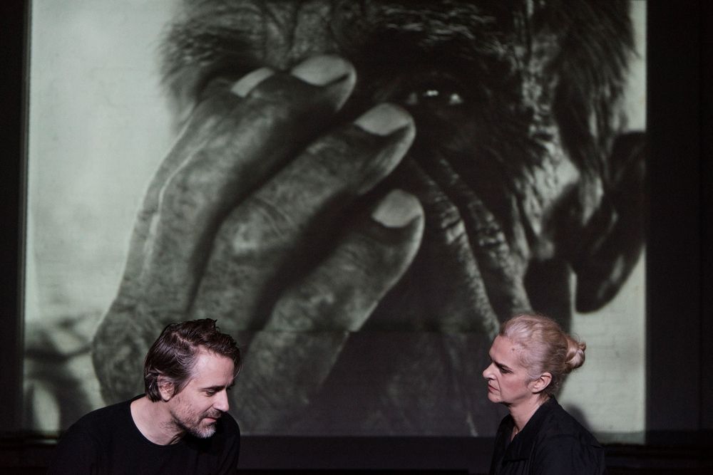 Szenenfoto mit zwei Personen, hinter ihnen eine große Schwarz-Weiß-Projektion eines Fotos einer Person, die beide Hände vors Gesicht hält