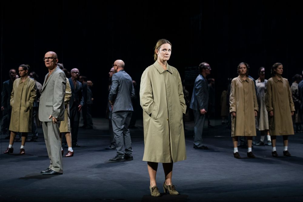 Szenenfoto "1984" mit mehreren Personen in Anzügen und Trenchcoat, im Vordergrund eine Frau mit Trenchcoat, die Menschen spiegel sich in zwei großen Spiegelfächen im Hinteren des Bildes