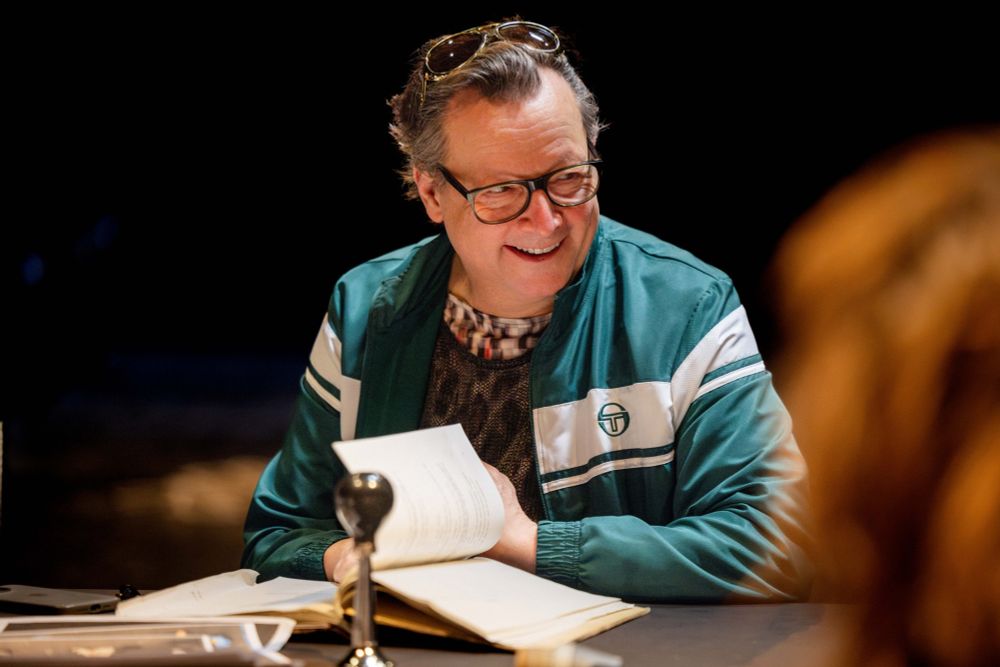 Matthias Brandt lachend im Probenkostüm mit Sportjacke und Sonnenbrille auf dem Kopf vor einem Textbuch am Tisch