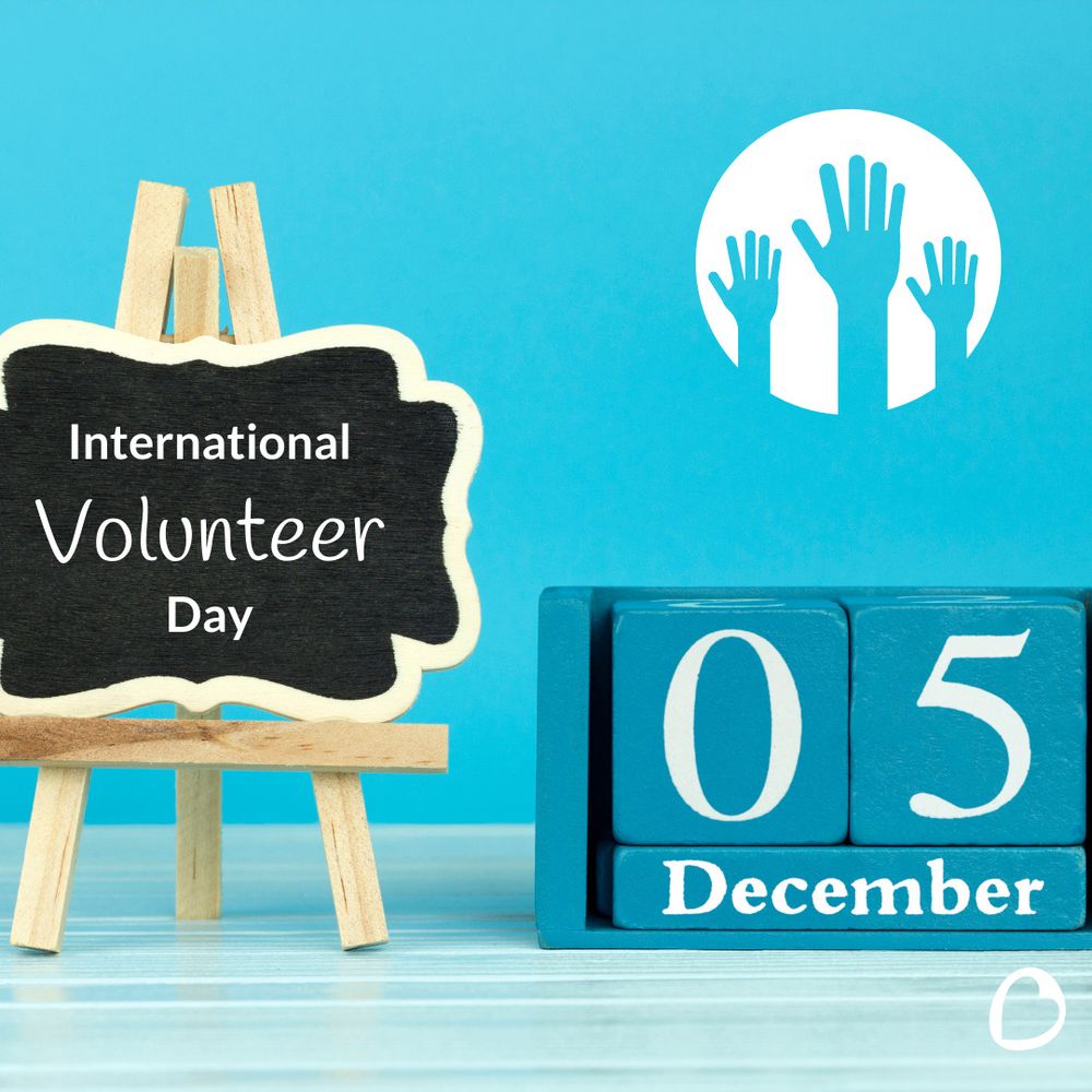 A small chalkboard on an easel reads "International Volunteer Day" against a blue background. Next to it, a block calendar displays "05 December." Above, a graphic shows three raised hands in a circle, symbolising volunteering.

