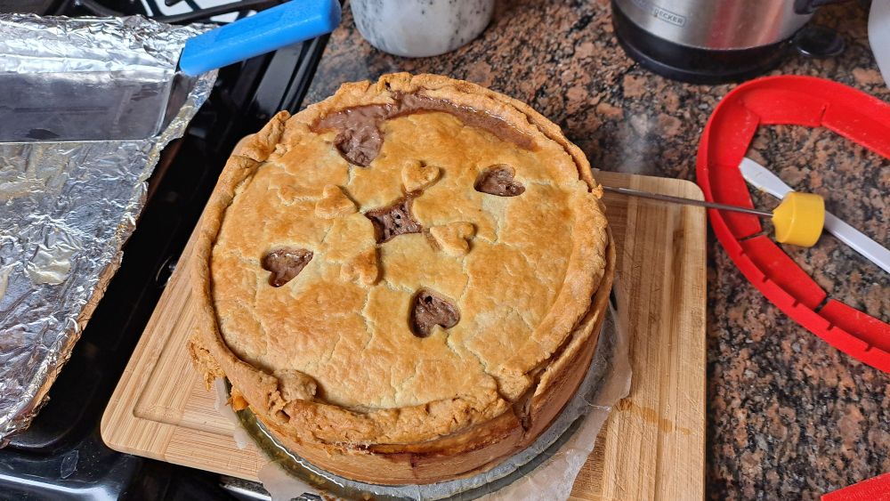 A tall meat pie with a full pastry coffin; 4 heart shapes and 1 four-pointed star are cut into the golden top crust