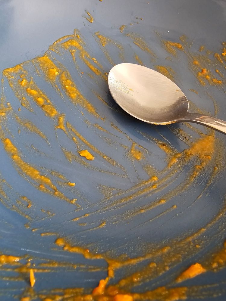 Ein benutzter blauer Suppenteller, in dem ein Löffel liegt. Auf dem Teller sind orangefarbene Essensreste