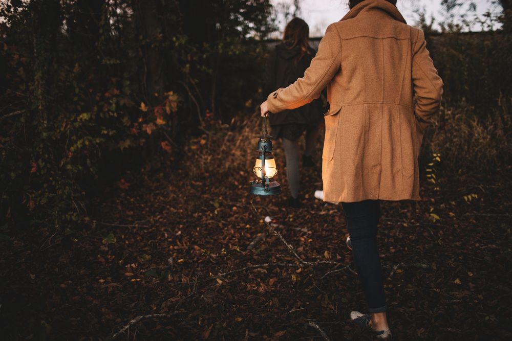 A stock photo of a woman wearing a tan coat and jeans, holding a lantern while walking through a dark forest with autumn leaves blanketing the ground around her.