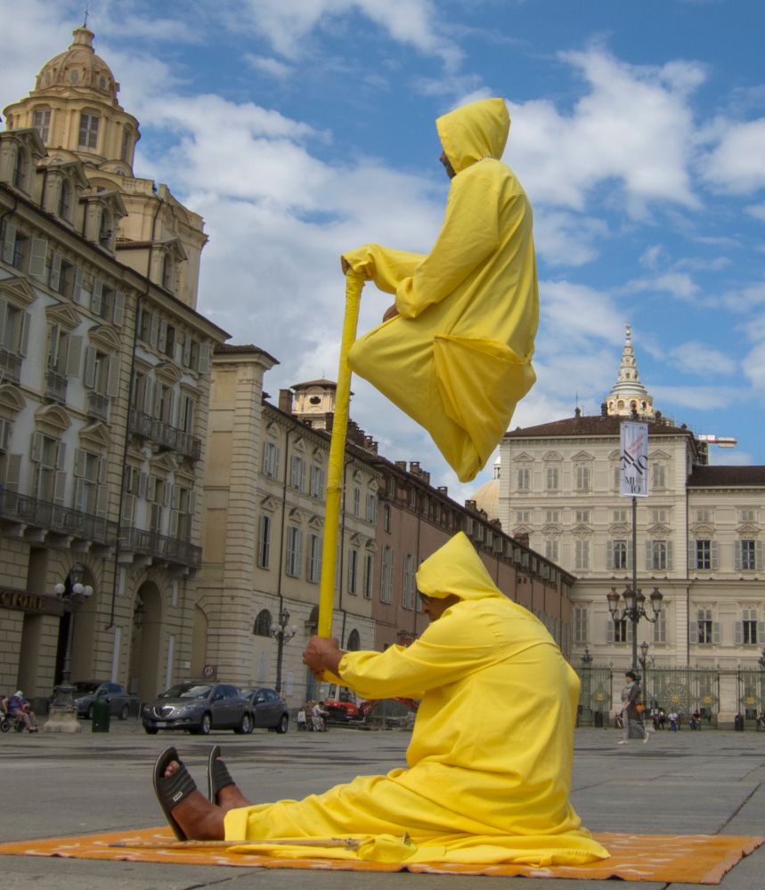 auf einem Stadtplatz sind zwei übereinandersitzende Personen in gelben Kostümen mit Kapuzen zu sehen, wobei der Eindruck ensteht, das die untere Person einen dünnen Stock hält, an dem sich die obere Person nur mit der Hand festhält. sie quasi in der Luft sitzt.