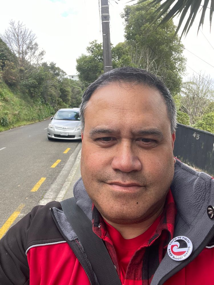 Matthew Reweti who is running for Wellington City Council’s Māori Ward, stands on a footpath. Behind is a typical Wellington suburban street with a car parked. In front of the car broken yellow line.
