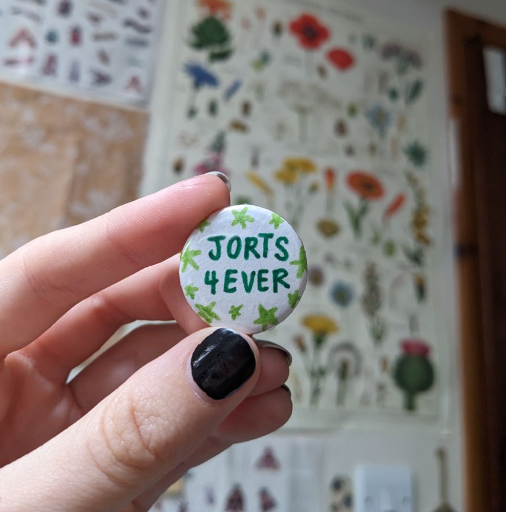 pin badge that says "jorts 4ever" in dark green on a white background with light green stars around the edges