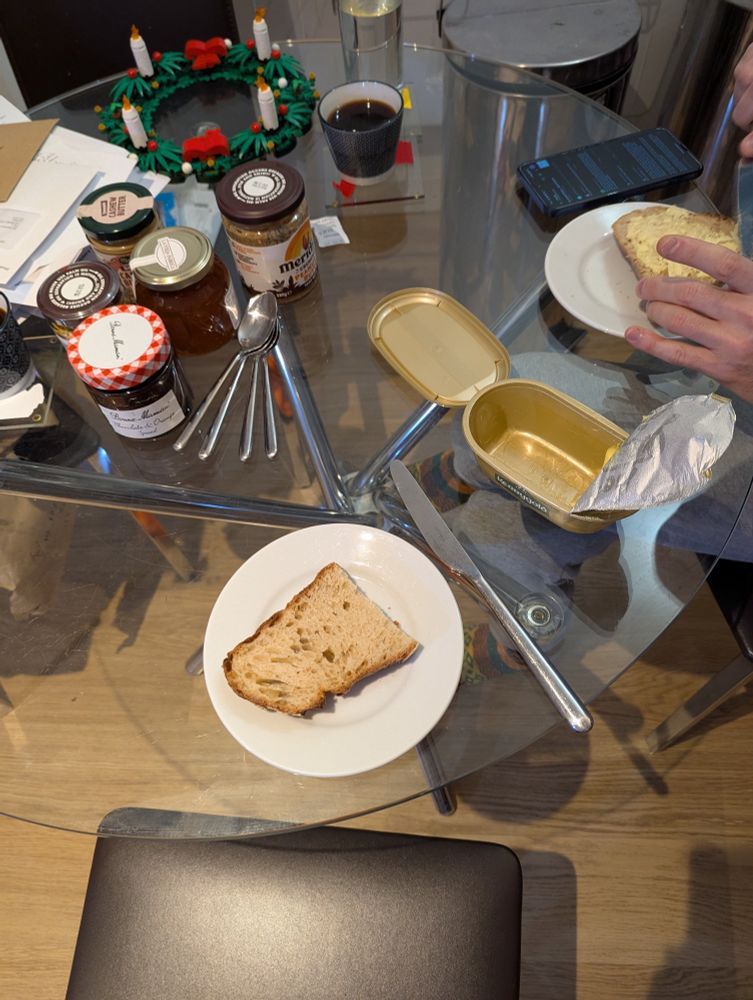 A glass kitchen table, with a fully-lit LEGO Advent wreath in the back, a stack of mail left background. The foreground contains two cups of coffee, a variety of jars of nut butter and jam, a pile of spoons, and two plates of toast at far right. The butter's between the two plates.