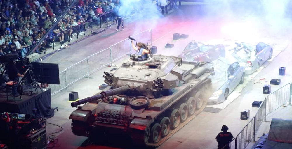 Foto aus einer großen Veranstaltungshalle. In der Bildmitte ein Panzer, der gerade vier Auto platt gewalzt hat, in der geöffneten Turmluke des Panzers steht ein Typ in einer Jeansweste und hält zwei AR-15 in die Luft. Hinter dem Panzer ist viel Nebel (vermutlich Nebelmaschinen und Abgase), in der oberen linken Ecke ist eine Tribüne mit Publikum zu erkennen
