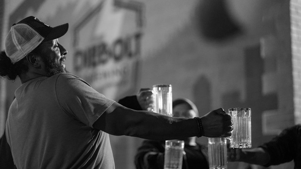 Black-and-white photo of a stein holding competitor at DPO’s Oktoberfest event. The person in the foreground wears a baseball cap and short-sleeved shirt, holding a heavy glass stein filled with liquid at arm’s length. Other participants are visible behind, all engaged in the endurance challenge. A mural with partial “DIEBOLT BREWING” text anchors the setting.