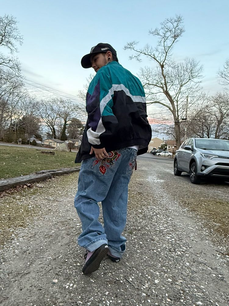 korri wearing a hat, vintage windbreaker, baggy embroidered jeans, and purple shoes. he is turned away from the camera.