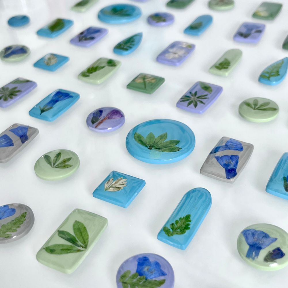The image shows polymer clay pieces laid out over a desk with a white background. The polymer clay pieces are organized in a nice grid pattern and are all different shapes, including circles, squares, rectangles, and arches. On the shapes there are pieces of different plants, including bluebells, ferns, and other leaves. They are coated in resin getting ready to be made into earrings.
