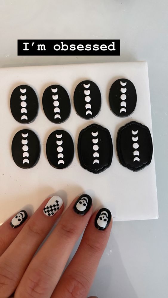 Hand with black and white nail polish with skulls and checkerboard details over black ovals that have white moon phases over them that are prepped polymer clay earrings