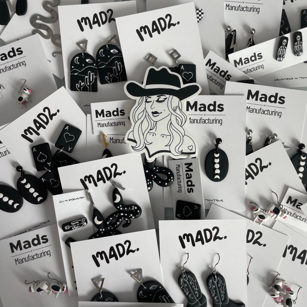 The photo is black and white earrings all over the desk. All of the earrings are on earring cards. The earrings are black, playing cards, black moon, phases, black and white snakes, black and white cows, and black and white cowboy boots. There is a picture of a goth cowgirl sticker on top of all of the earrings. 