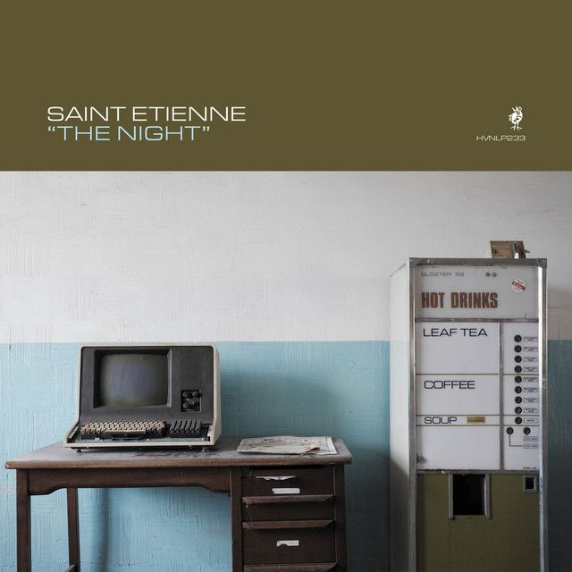 The cover of Saint Etienne's album "The Night." It shows an old, all-in-one computer on an equally old desk. Next to it is an even older HOT DRINKS machine which dispenses leaf tea, coffee, and soup. Behind both is a wall, the upper part of which is painted white and the lower part of which is painted in a muted light blue. The overall mood is one of times gone by and obsolescence.