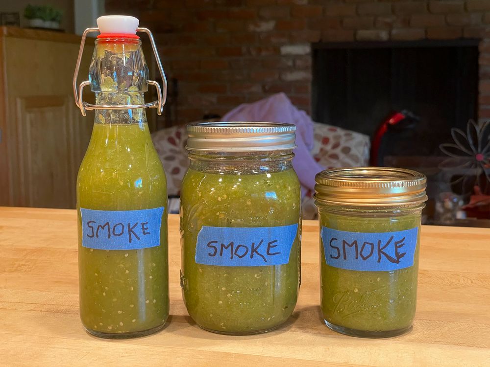 Homemade green jalapeno hot sauce in jars on tabletop. Two are canning jars (12oz. and 16oz.) and one is a swing top w/red rubber seal. Shot in my living room showinh clutter behind the jars.
