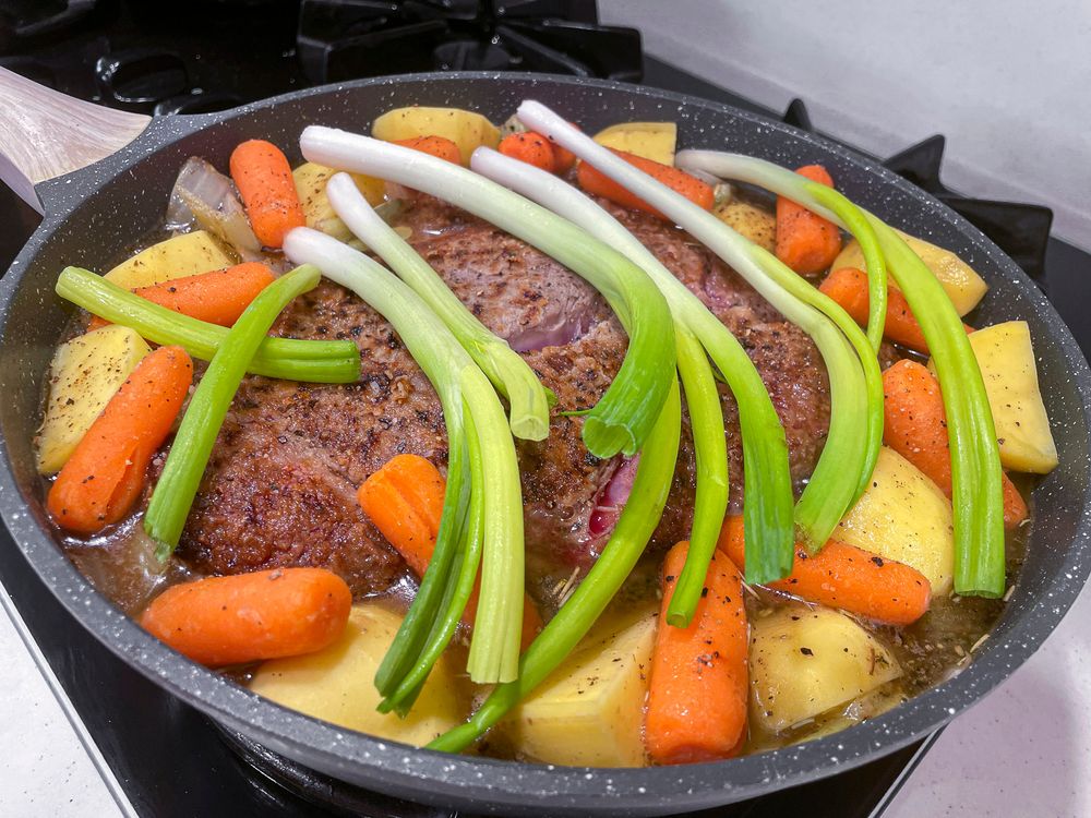 Large pan on the stove top holding a small browned beef chuck roast in the center, surrounded by onions, carrots and potatoes on the sides, and topped with green onions. Celery (that you can't see) is on the bottom underneath everything.