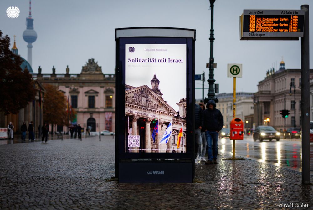 Eine Werbetafel an einer Bushaltestelle in Berlin-Mitte zeigt das Kampagnenbild des Deutschen Bundestages. Darauf zu sehen ist das Reichstagsgebäude, vor dem die europäische, die israelische und die deutsche Flaggen wehen. Außerdem ist zu lesen: "Solidarität mit Israel. Der Deutsche Bundestag steht solidarisch und entschlossen an der Seite Israels und seiner Menschen." Im Hintergrund der Bushaltestelle sieht man den Fernsehturm, Passanten und Verkehr.