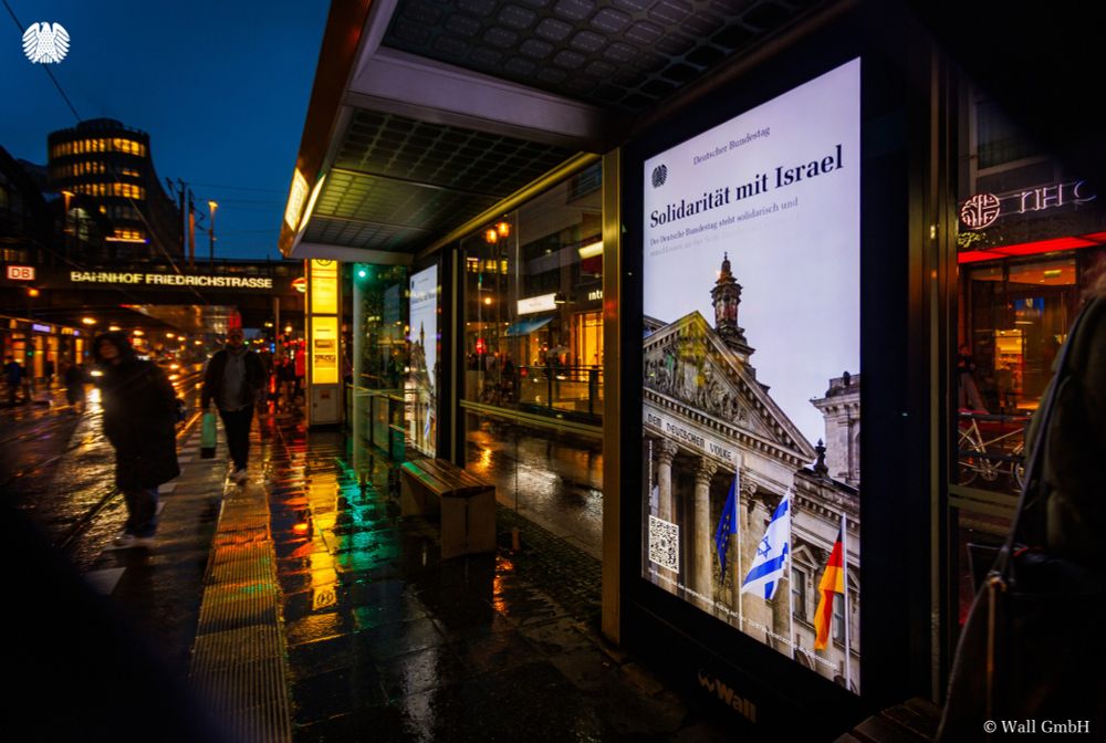 Eine Werbetafel an einer Haltestelle in Berlin-Mitte zeigt das Kampagnenbild des Deutschen Bundestages. Darauf zu sehen ist das Reichstagsgebäude, vor dem die europäische, die israelische und die deutsche Flaggen wehen. Außerdem ist zu lesen: "Solidarität mit Israel. Der Deutsche Bundestag steht solidarisch und entschlossen an der Seite Israels und seiner Menschen." Im Hintergrund sieht man den S-Bahnhof Friedrichstraße, Passanten und Verkehr.