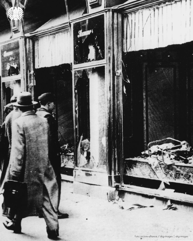 Das Foto zeigt ein in der Reichspogromnacht durch die Nationalsozialisten zerstörtes jüdisches Geschäft. Auf der Straße vor dem zerstörten Geschäft geht ein Mann vorbei, andere schauen sich die Zerstörung an.