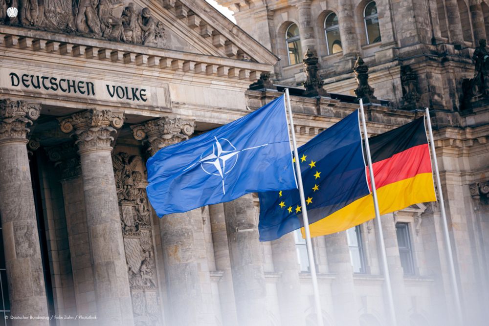 Vor dem Reichstagsgebäude weht die Flagge der NATO neben der Europaflagge und der Bundesflagge (von links nach rechts). Die Flaggen sind im Vordergrund an Masten befestigt; im Hintergrund sieht man Teile des Reichstagsgebäudes.