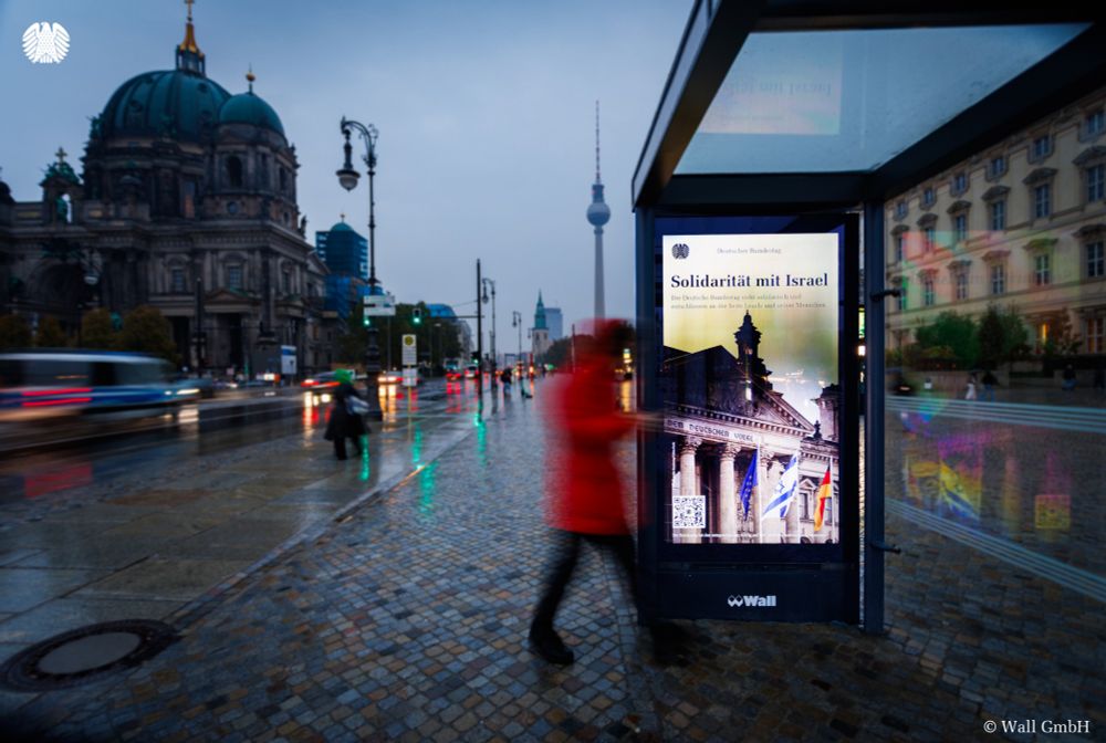 Eine Werbetafel an einer Bushaltestelle in Berlin-Mitte zeigt das Kampagnenbild des Deutschen Bundestages. Darauf zu sehen ist das Reichstagsgebäude, vor dem die europäische, die israelische und die deutsche Flaggen wehen. Außerdem ist zu lesen: "Solidarität mit Israel. Der Deutsche Bundestag steht solidarisch und entschlossen an der Seite Israels und seiner Menschen." Im Hintergrund der Bushaltestelle sieht man den Berliner Fernsehturm, den Dom, Passanten und Verkehr.
