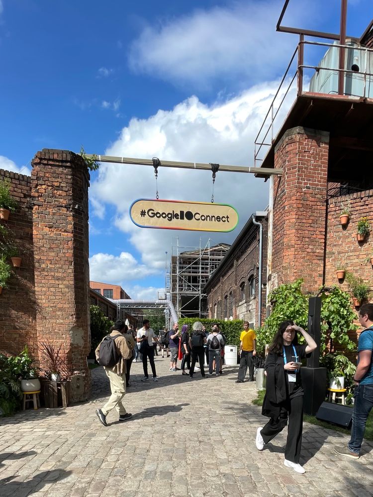 A industrial yard with red bricks, a wooden #GoogleIOConnect sign, and plants and people wandering around.