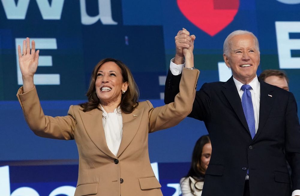 A joyful President Biden and Vice President Harris link hands and wave at an event.