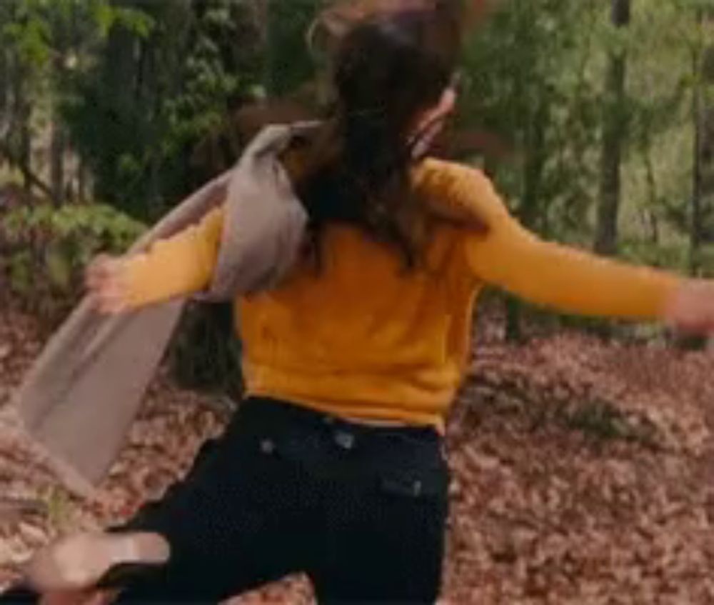 a woman in a yellow sweater and black pants is running through the woods with her arms outstretched .