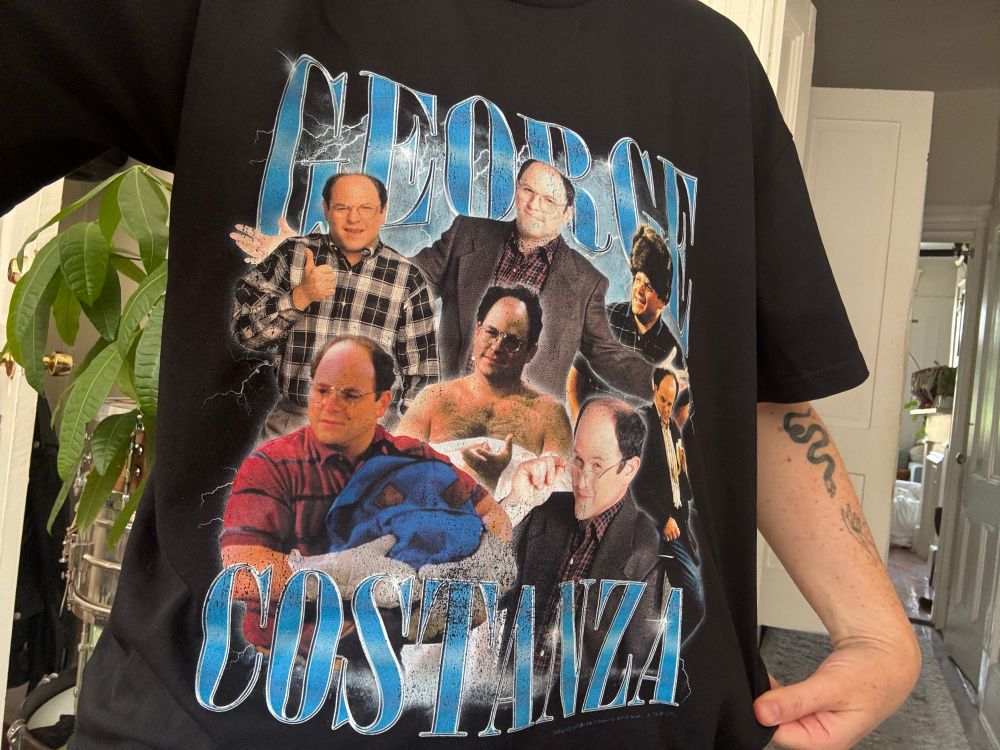 Black tee shirt with various pictures of actor Jason Alexander from Seinfeld against a lightening background with the words GEORGE COSTANZA in blue. 
