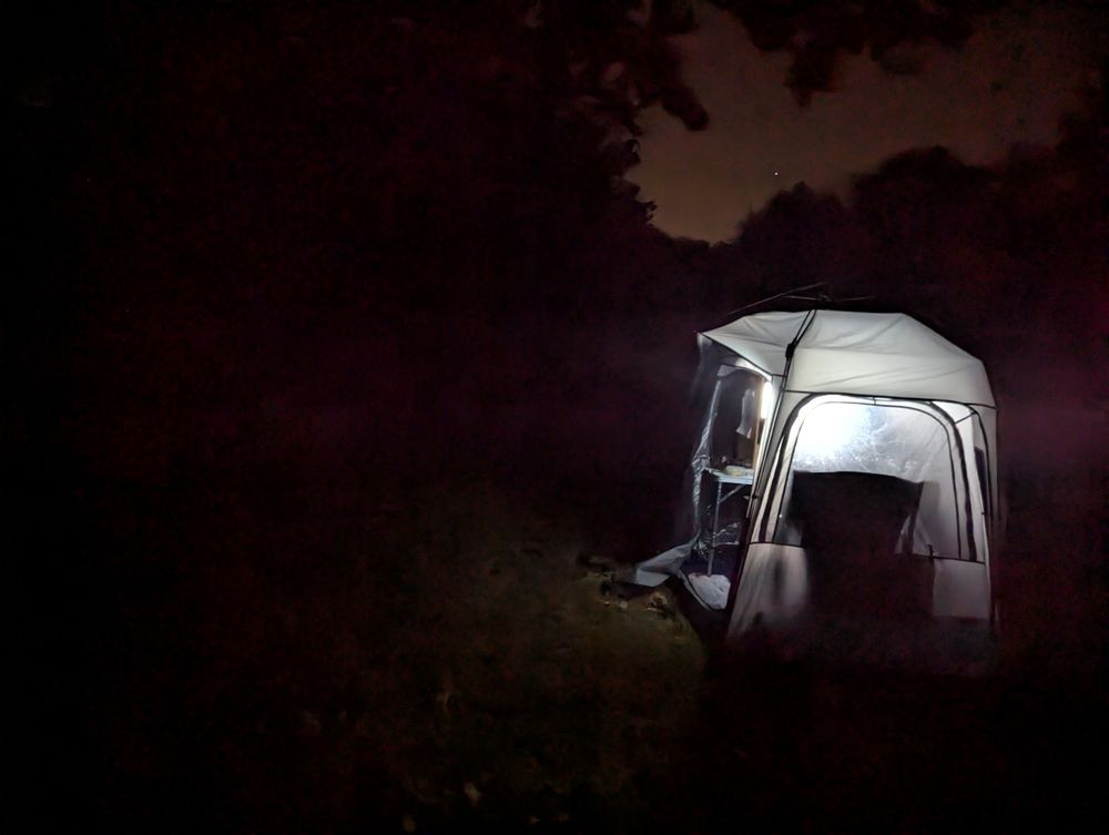 Looking east along a two-lane highway.
It is nighttime and there are no street lights.
In the foreground is the ham pod.
The inside the ham pod is illuminated by a computer screen, giving the pod an eerie glow.
A chair inside the pod casts a dark shadow.
The sky is only barely lighter than the surroundings, and the silhouettes of trees frame a single bright star.
The grass immediately left of the ham pod is faintly lit.