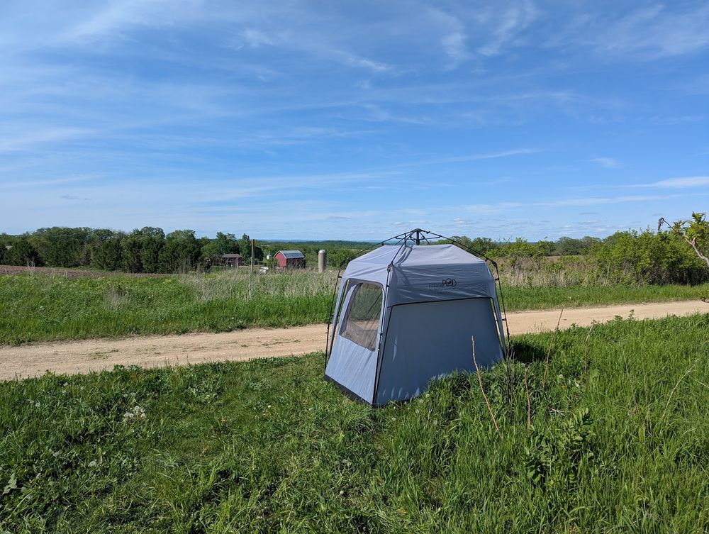 Looking north across a State Trail.
The trail is light brown dirt and gravel, a "Strada Bianca".
The trail is lined with grasses along both sides.
In the grass on the near side of the trail is the grey ham pod.
The sky has quite a few wispy clouds, but otherwise appears blue.
There are some farm buildings in the distance.