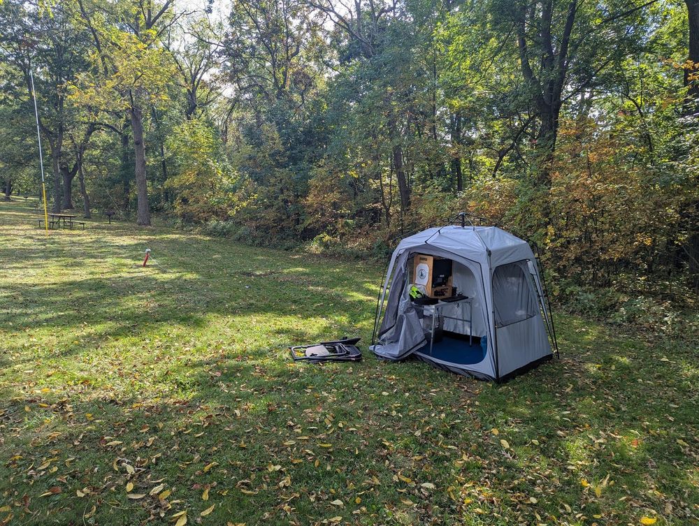 A grassy field surrounded by trees.
To the right is the ham pod.
Visible inside the ham pod are a small table under which sits a large silver battery.
Atop the table are a radio box, a computer keyboard, and some gloves.
To the left of the ham pod, a folding chair lies in the grass.
A red post is visible, with an antenna strung from it to a yellow pole at the left, next to a picnic table.
A few cookout grills are visible near the trees.