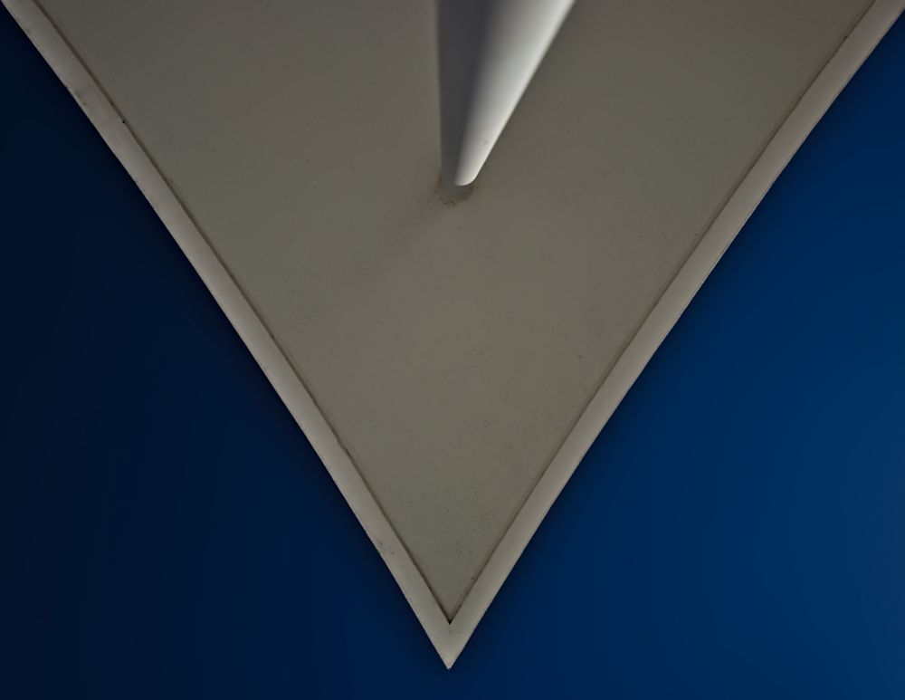 Das Bild zeigt die Unterseite eines Daches, das aus einer ungewöhnlichen Perspektive fotografiert wurde. Die dreieckige Form des Daches ragt in das Bild hinein, mit klar definierten Kanten und einer dezenten Schattenwirkung. Die helle, beige oder graue Oberfläche des Daches steht in starkem Kontrast zum tiefblauen Hintergrund, wodurch die rechteckige Form besonders hervorgehoben wird.