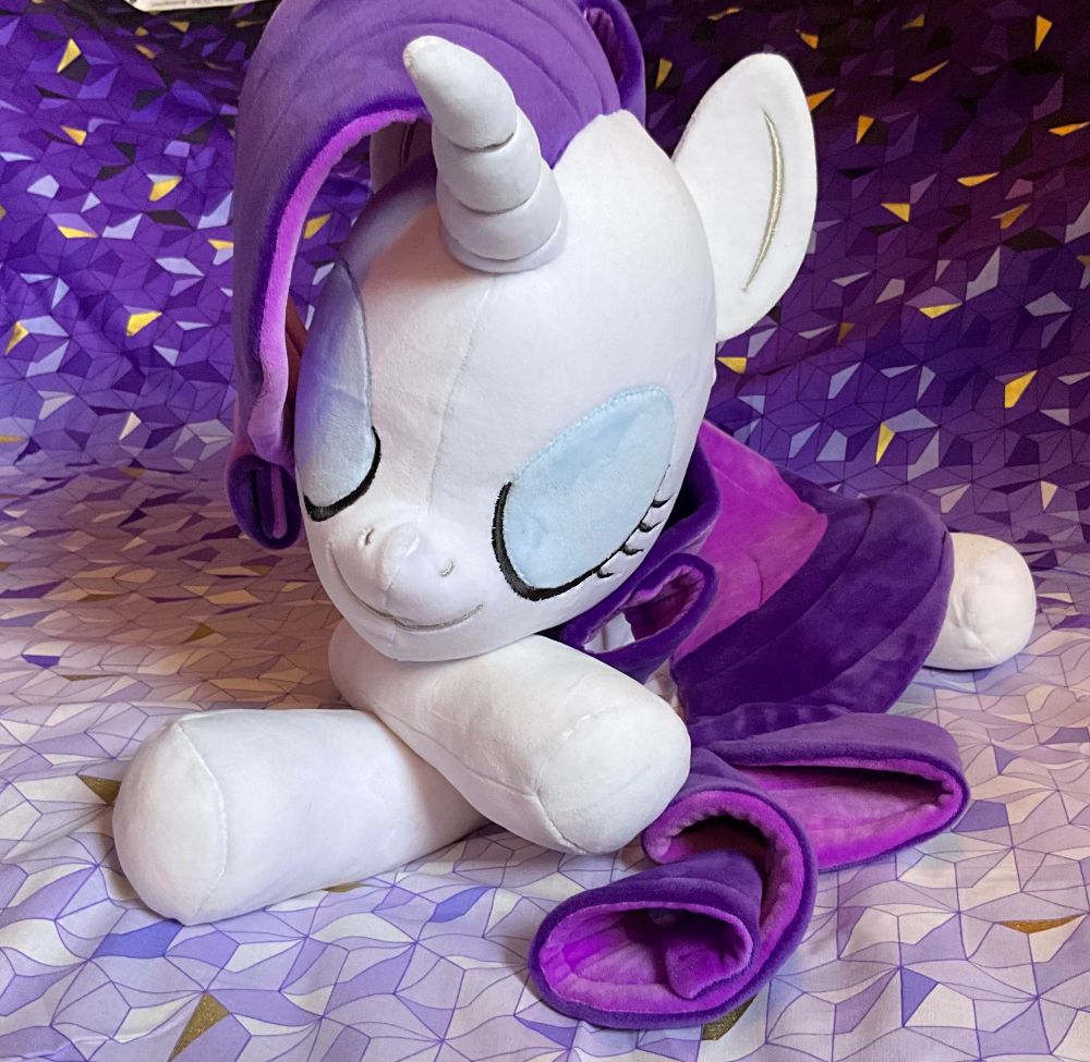 A photo of a handmade 20-inch plush doll of Rarity from My Little Pony: Friendship is Magic in a laying pose with a relaxed, sleeping expression against a purple backdrop as seen from the front.