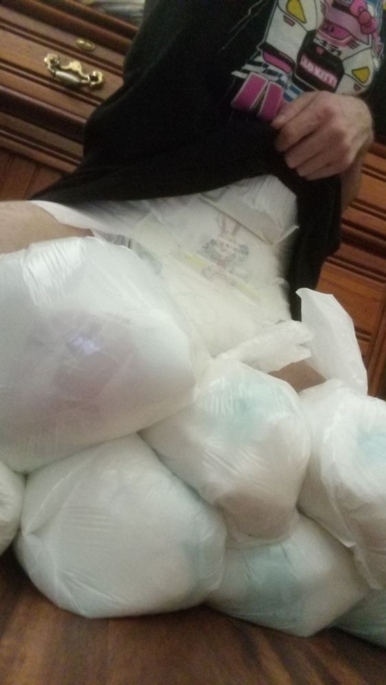 I am sitting on a pile of big, used diapers tied up in small diaper bags. There are 7 below me. I am wearing pink thigh high socks and a black Hello Kitty shirt. I am lifting up my shirt to show you my diaper .