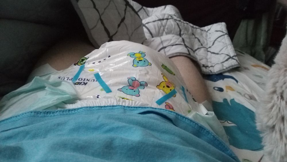 Me tenting my Kiddo Junior Maxi Plus diaper. Just woke up. Wearing a blue shirt. Can see a bit of my dino bed sheets and my bunny plushy. Towel in the background in the right corner.