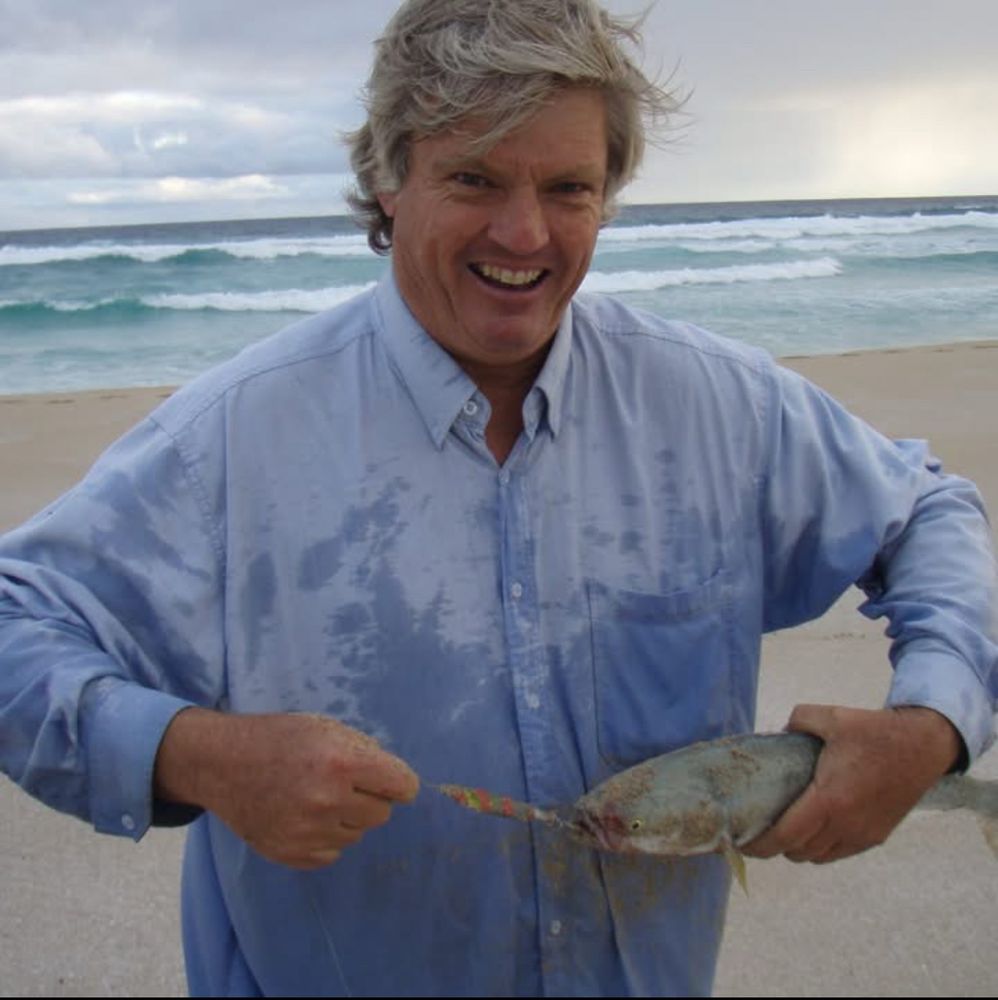 An unflattering picture of an old white man, Stephen Heatley, standing on a beach holding a fish and smiling.