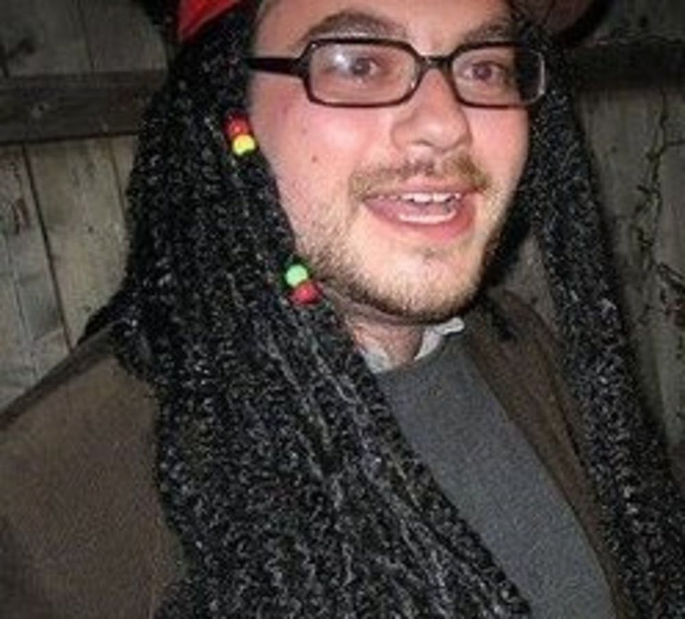 Matt Yglesias, a chubby white man with glasses and a scruffy goatee, is wearing a racist Jamaican braid wig, with his mouth open in a stupid expression.