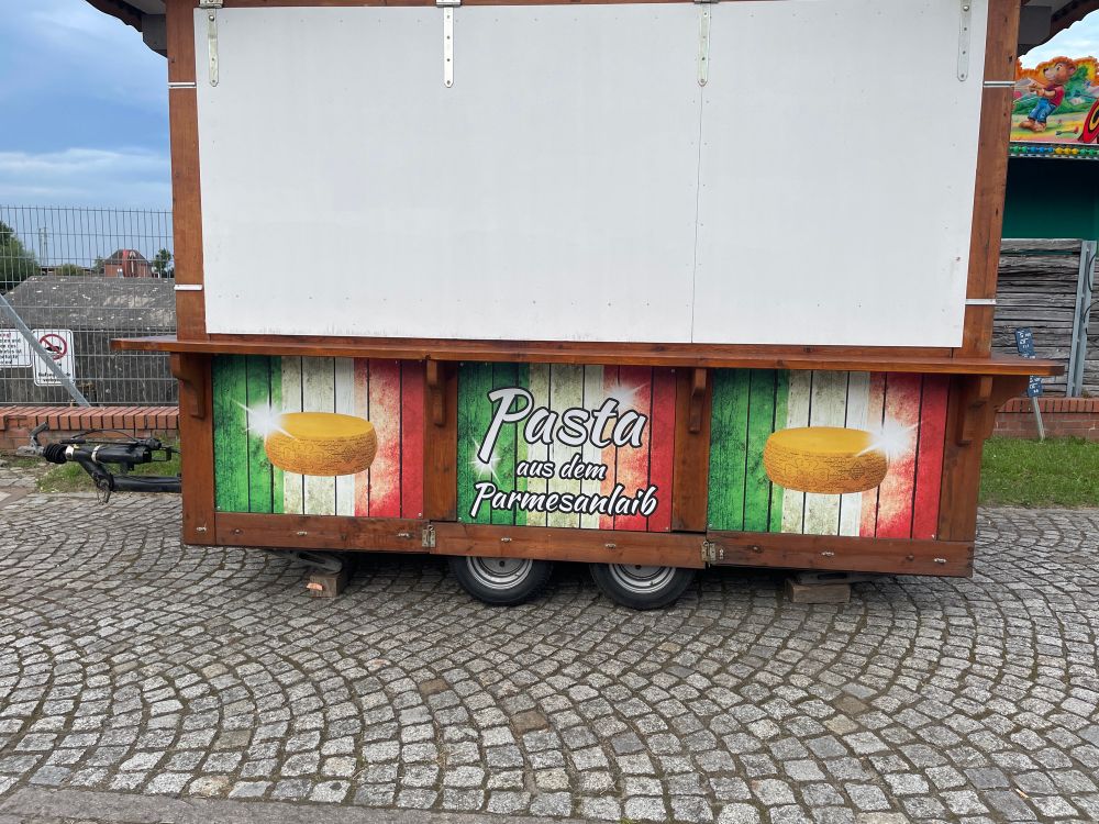 Foto eines (gerade geschlossenen) Jahrmarkt-Verkaufstand, Aufschrift: „Pasta aus dem Parmesanlaib“, links und rechts daneben sind Parmesanlaibe abgebildet 