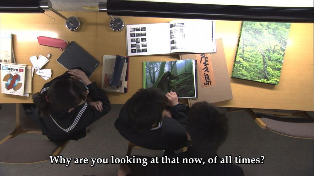 Asumu and friends studying in school. One of them asks: "Why are you looking at that now, of all times?" (He's staring down at a textbook with a picture of a forest)